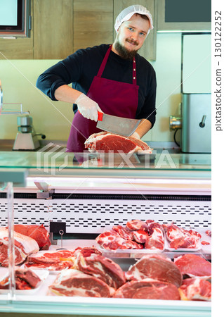 Young bearded butcher slicing fresh beef for customer at counter 130122252