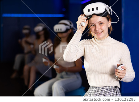 Cheerful young girl in VR headset on head with joysticks in hands looking with excited expression 130122293