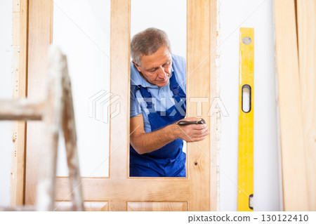 Professional middle-aged repairman in uniform installing a door handle on a door during renovation, indoors Professional middle-aged repairman in uniform installing a door handle on a door during renovation, indoors 130122410