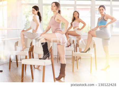 Leaning on chair seat with hand foot, young woman performs lap dance during group workout. 130122428