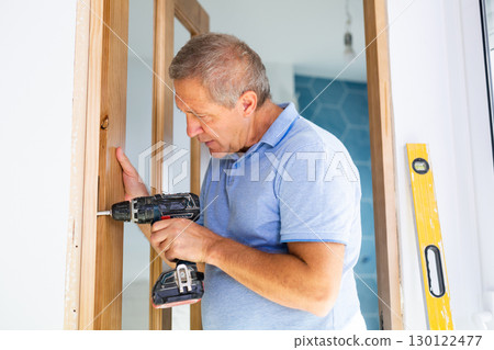 Mature man himself installs the door hinges with screwdriver 130122477