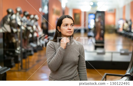 Interested female tourist visiting exhibition of medieval armor in museum 130122491
