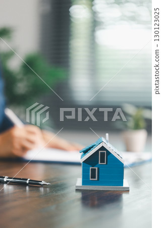 Real estate agent and customer are discussing contract to buy house, with house model in background symbolizing real estate, insurance, and loan arrangements involved in process. 130123025