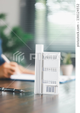 Real estate agent and customer are discussing contract to buy house, with house model in background symbolizing real estate, insurance, and loan arrangements involved in process. Real estate agent and customer are discussing contract to buy house, with house model in background symbolizing real estate, insurance, and loan arrangements involved in process. 130124552