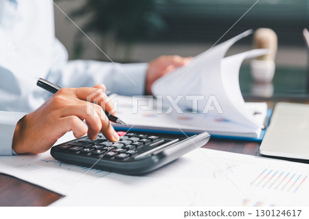 Office businesswoman hand to operate calculator analyzing financial data, counting costs on desk. accounting, financial analysis for business budget planning, calculation support, finance management. Office businesswoman hand to operate calculator analyzing financial data, counting costs on desk. accounting, financial analysis for business budget planning, calculation support, finance management. 130124617