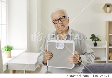 Upset senior man holding scales with frustrated face standing in the living room at home. 130124671