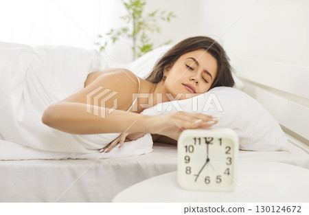 Peaceful young woman sleeping comfortably in bed with alarm clock nearby, resting early in morning. 130124672