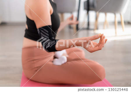 Close up view of middle aged woman sitting cross legged in lotus pose on pink yoga mat in modern apartment, focusing on yoga and flexibility 130125448