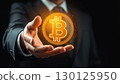A futuristic image of a businessman in a suit holding a glowing holographic Bitcoin symbol in his hand, surrounded by digital icons and data points. 130125950