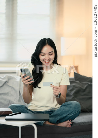 Smiling young woman holding a credit card and smartphone while shopping online at home 130127408