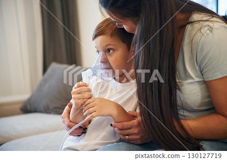 Boy is making inhalation with nebulizer to reduce coughing. Mother with her little son is at home in domestic room Boy is making inhalation with nebulizer to reduce coughing. Mother with her little son is at home in domestic room 130127719