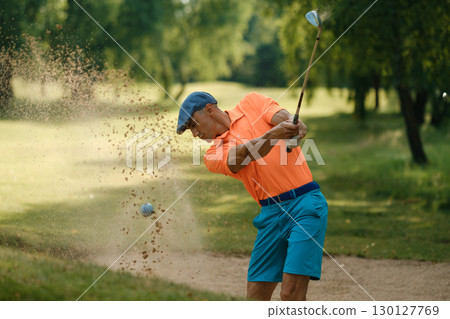 Golf player makes a powerful swing from a sand trap on a sunny day at a lush green golf course surrounded by trees Golf player makes a powerful swing from a sand trap on a sunny day at a lush green golf course surrounded by trees 130127769
