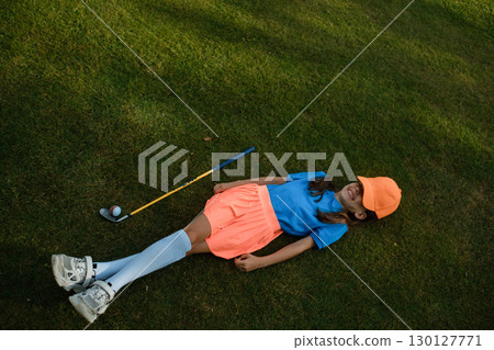 A young girl in a bright sports outfit is relaxing on the green grass on a sunny day at the golf course, tired after half a workout A young girl in a bright sports outfit is relaxing on the green grass on a sunny day at the golf course, tired after half a workout 130127771