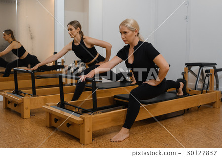 Two women perform Pilates exercises in a studio with wooden reformers, focusing on flexibility and strength training during a morning session 130127837