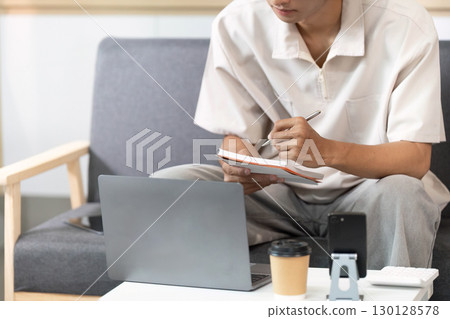 Note-Taking. Young man writing in notebook while using laptop at home. 130128578