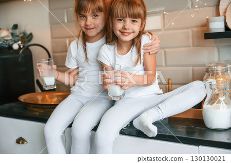 Embracing each other. Happy two little girls are drinking milk while sitting on the table on the kitchen Embracing each other. Happy two little girls are drinking milk while sitting on the table on the kitchen 130130021