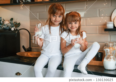 Embracing each other. Happy two little girls are drinking milk while sitting on the table on the kitchen Embracing each other. Happy two little girls are drinking milk while sitting on the table on the kitchen 130130025