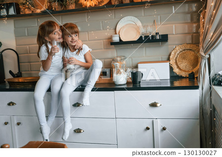 Happy two little girls are drinking milk while sitting on the table on the kitchen Happy two little girls are drinking milk while sitting on the table on the kitchen 130130027