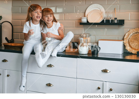 Wellness of the people. Happy two little girls are drinking milk while sitting on the table on the kitchen Wellness of the people. Happy two little girls are drinking milk while sitting on the table on the kitchen 130130030