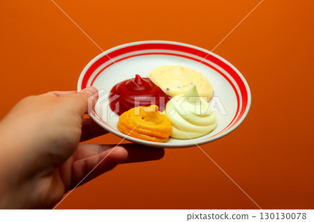 a hand holding a plate with a variety of sauces, including ketchup, mustard, mayonnaise, and garlic sauce on dark orange background. hand holding dipping sauces plate a hand holding a plate with a variety of sauces, including ketchup, mustard, mayonnaise, and garlic sauce on dark orange background. hand holding dipping sauces plate 130130078