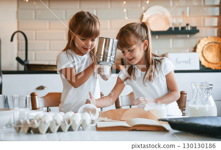 Using flour. Two little girls are preparing food on the kitchen with garlands Using flour. Two little girls are preparing food on the kitchen with garlands 130130186