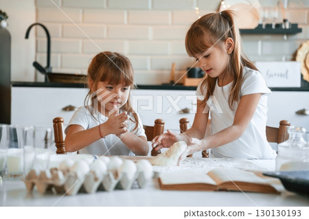Flour and eggs on the table. Two little girls are preparing food on the kitchen with garlands Flour and eggs on the table. Two little girls are preparing food on the kitchen with garlands 130130193
