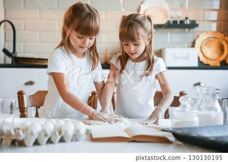 Flour and eggs on the table. Two little girls are preparing food on the kitchen with garlands Flour and eggs on the table. Two little girls are preparing food on the kitchen with garlands 130130195