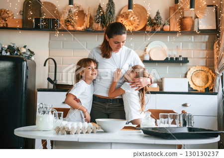 Mother with her little two daughters is preparing food on the kitchen with garlands together Mother with her little two daughters is preparing food on the kitchen with garlands together 130130237