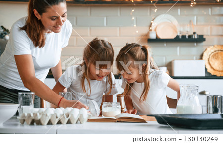 Using quality products. Mother with her little two daughters is preparing food on the kitchen with garlands together Using quality products. Mother with her little two daughters is preparing food on the kitchen with garlands together 130130249