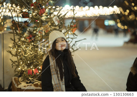 Asian smiling girl in white hat sitting in front of Christmas tree with lights and holiday decorations, outdoor night scene with bokeh, winter joy, holidays, childhood memories 130130663