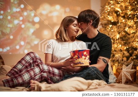 With popcorn. Beautiful young couple indoors, celebrating new year against projector 130131177