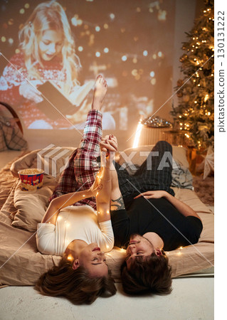 Lying down on bed, holding hands. Beautiful young couple indoors, celebrating new year against projector 130131222