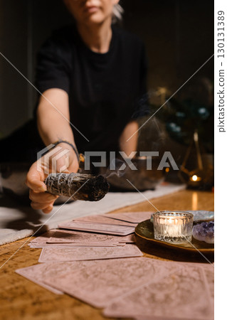 Woman performing a cleansing ritual with sage near a candle and crystal on a wooden table surrounded by plants in an intimate indoor setting 130131389