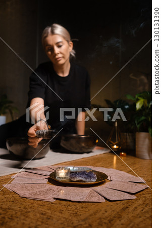 Woman performing a cleansing ritual with sage near a candle and crystal on a wooden table surrounded by plants in an intimate indoor setting 130131390
