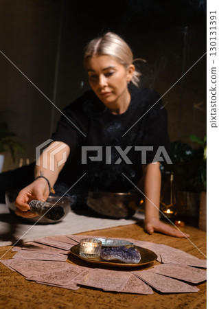 Woman performing a cleansing ritual with sage near a candle and crystal on a wooden table surrounded by plants in an intimate indoor setting 130131391