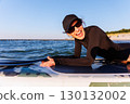 Smiling woman enjoys surfing on Baltic Sea waves, laughing joyfully lying on board with splashing water around. Bright sunlight and her laughter reflect pure joy, freedom and vibrant summer adventure 130132002