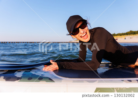 Smiling woman enjoys surfing on Baltic Sea waves, laughing joyfully lying on board with splashing water around. Bright sunlight and her laughter reflect pure joy, freedom and vibrant summer adventure 130132002