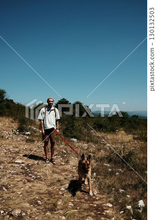 Man hiking with German Shepherd on rocky mountain trail in Stara Planina, Serbia, under bright blue summer sky 130132503