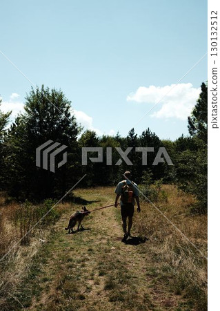 Man with backpack hiking with his German shepherd dog on forest trail in Stara Planina in Serbia country under summer sunlight. Rear view 130132512
