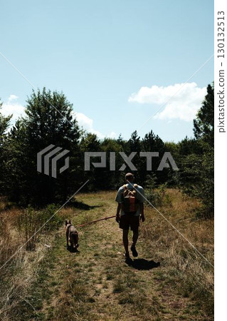 Man with backpack hiking with his German shepherd dog on forest trail in Stara Planina in Serbia country under summer sunlight. Rear view 130132513