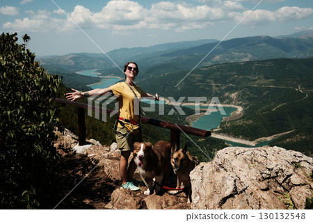 Woman spreading arms in joy with dogs German and Australian Shepherd on mountain viewpoint, stunning turquoise river winding through valley below. Travel and hiking with pets concept. Woman spreading arms in joy with dogs German and Australian Shepherd on mountain viewpoint, stunning turquoise river winding through valley below. Travel and hiking with pets concept. 130132548