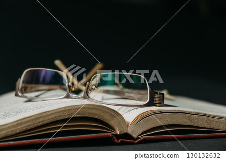 open book resting on a table with eyeglasses beside it in dimly lit room. closeup. 130132632