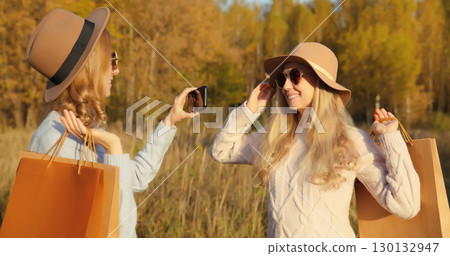 Happy women friends taking selfie with phone, shopping bags in autumn, girlfriends smiling in park Happy women friends taking selfie with phone, shopping bags in autumn, girlfriends smiling in park 130132947