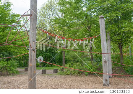A wooden playground structure with red ropes and metal rings set against a lush green park background, offering an adventurous climbing experience for children. A wooden playground structure with red ropes and metal rings set against a lush green park background, offering an adventurous climbing experience for children. 130133333