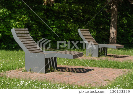 Two modern chaise lounges sit empty in a serene park setting. The chairs are positioned on brick tiles surrounded by lush greenery, offering a peaceful and inviting atmosphere for relaxation. Two modern chaise lounges sit empty in a serene park setting. The chairs are positioned on brick tiles surrounded by lush greenery, offering a peaceful and inviting atmosphere for relaxation. 130133339