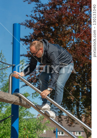 Man in jeans and jacket climbs a modern playground setup. Sunny weather, outdoor fitness, and a relaxed lifestyle moment for adult recreation in urban surroundings. 130133340