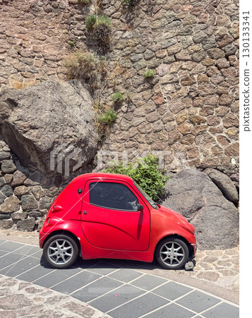 Red compact car parked against a textured stone wall in an urban setting. The vibrant red color contrasts with the natural stone backdrop, creating a unique visual scene. 130133341