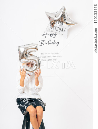 Cheerful five-year-old boy in white shirt posing with silver balloon on birthday. Ideal for childrens events, birthday cards, family content, and editorial or advertising use 130133358