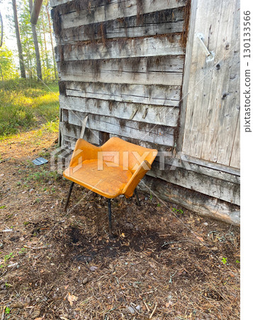 Worn yellow chair with metal legs leaning against aged wooden wall, surrounded by forest floor 130133566