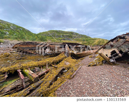 Decaying wooden shipwreck remains on rocky shore under cloudy mountain sky. 130133569
